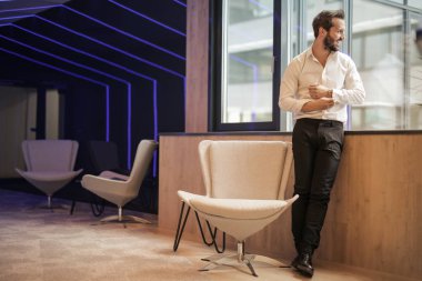 Kendine güvenen genç işadamı standları ve ofisinde pencereden görünüyor. 