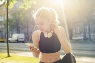 Sarışın genç kadın fitness kıyafetiyle şehirde onun smartphone kontrol kulaklıklar ile.