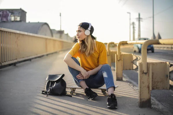 Young blonde teenager with baseball hat sitting on a bridge on her skateboard.