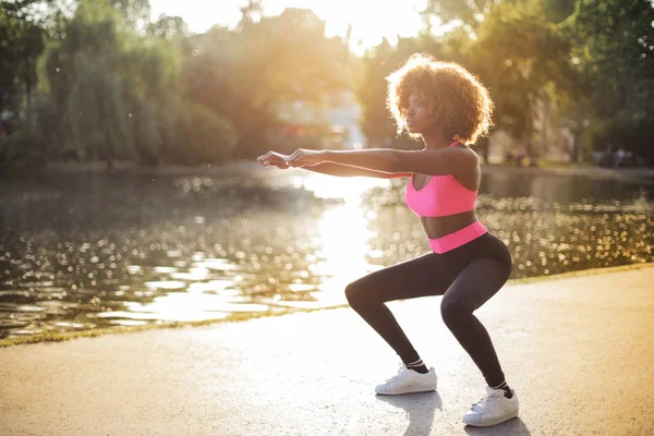 Güneşli bir günde bir göl yanındaki bodur egzersizleri yapıyor genç Afro kadın.