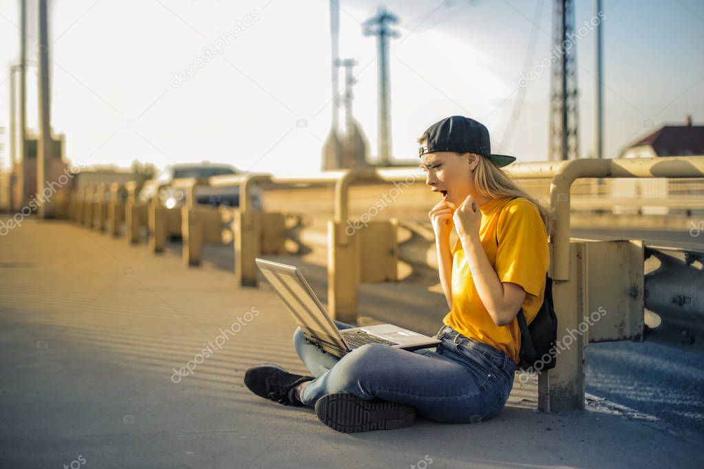 Young blonde woman sitting on a bridge in the city and happily checking her laptop.