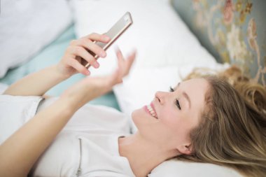 Onun yatakta yatan ve mutlu bir şekilde onun Smartphone cep telefonu ile internette sörf genç güzel sarışın kadın.