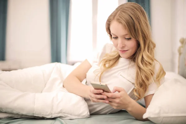 年轻的金发女人躺在她的床上 网上冲浪她的智能手机 — 图库照片