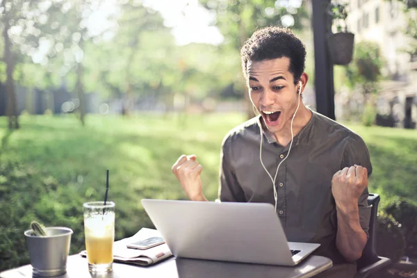 Young Afro man sitting on a terrace with a drink, watching his laptop gladly and feeling excited.