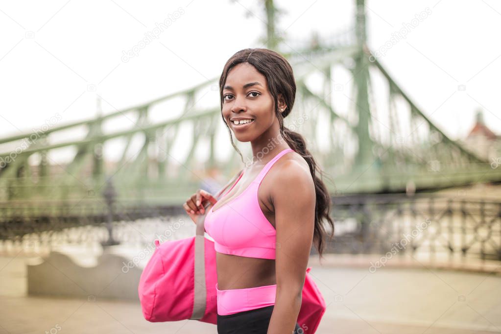 Young beautiful Afro woman in fitness outfit and a duffel bag walking on the street in the city.