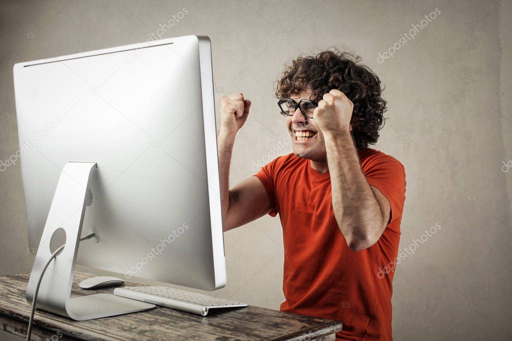 Young curly-haired man in front of a modern computer feeling extremely excited and happy.