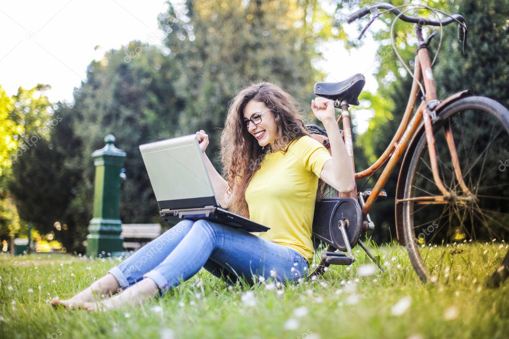Young curly-haired woman sitting in a park with her laptop and bike and feeling extremely excited.