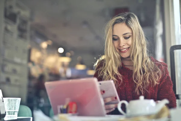 美丽的金发女孩与笔记本电脑检查她的智能手机在一个小酒馆和微笑 — 图库照片