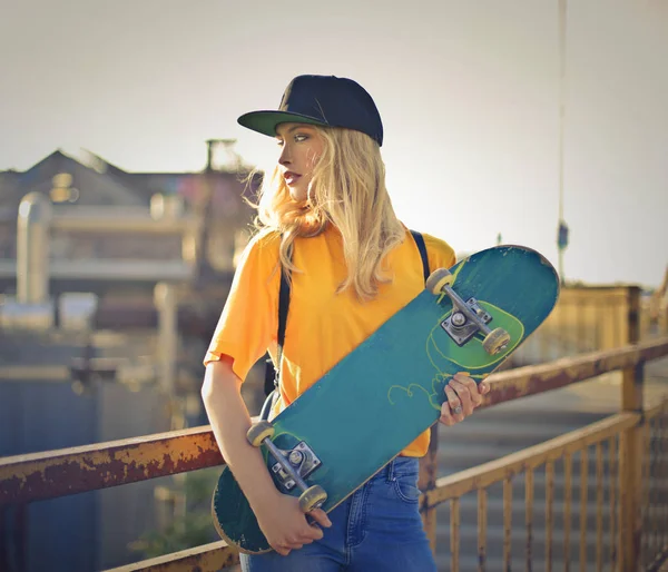 Young blonde woman with baseball hat and skateboard standing on a bridge in the city.