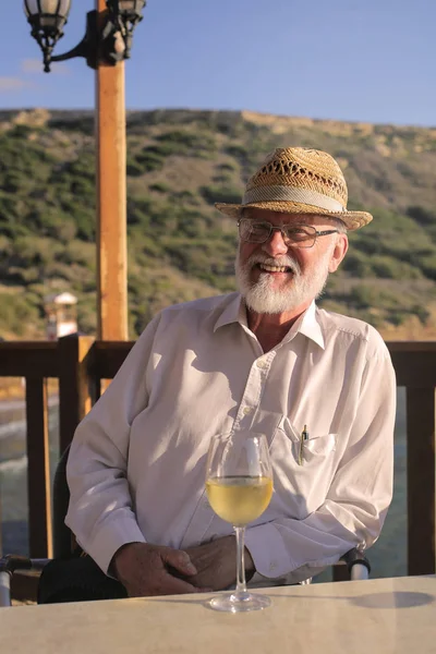 Grandfather with white beard, a straw hat and a glass of wine smiling positively on a terrace on a summer day.