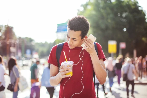Young Afro man with earphones and a drink walking on the street on a sunny day.