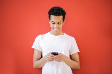 Onun smartphone kontrol ve kırmızı bir arka plan önünde gülümseyen genç Afro adam.