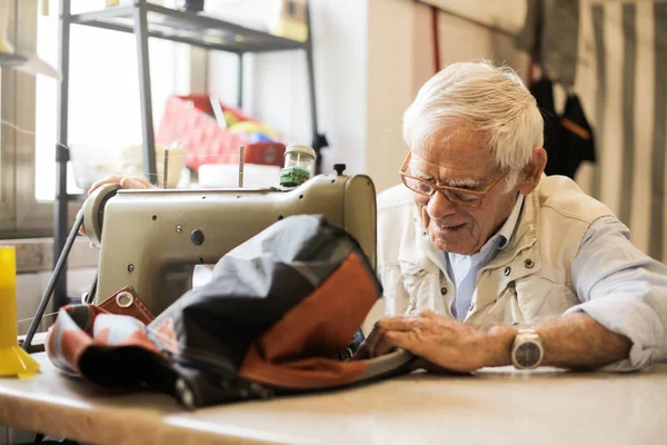 Elderly man with glasses sewing with a sewing machine in a workshop.