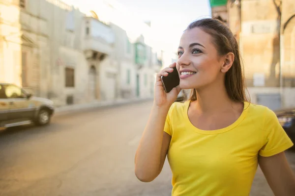 年轻美丽的女人穿着黄色 T恤走路和谈论在城市的智能手机 — 图库照片