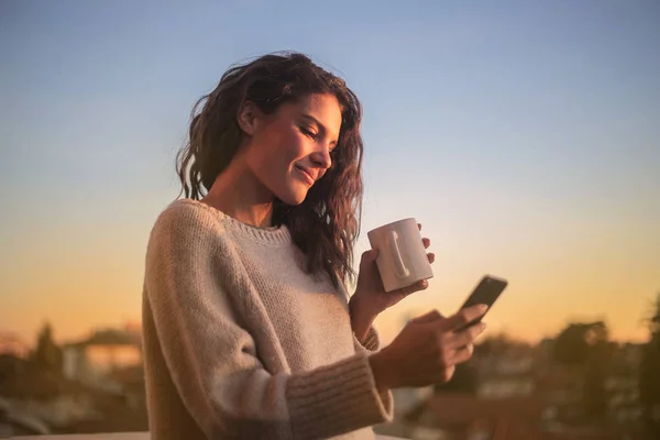 Young woman in the sunset checking her smartphone and drinking coffee.