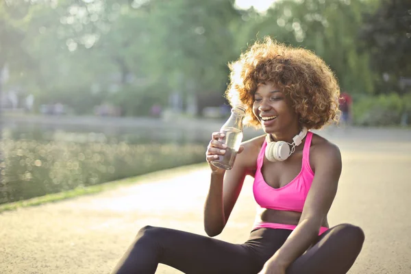 Young happy African woman taking a break during sport activity and drinking water in a park.