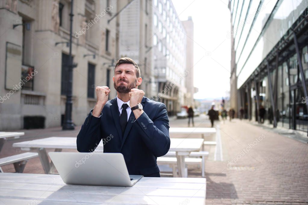 Young businessman with his laptop shows winner gesture in the city.