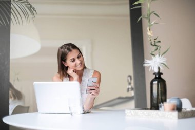 Bir dizüstü bilgisayar ile bir masada oturan ve onun smartphone kontrol genç kadın.