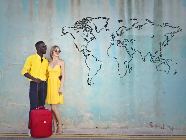 Afro man and blonde woman standing in front of a wall with a luggage.