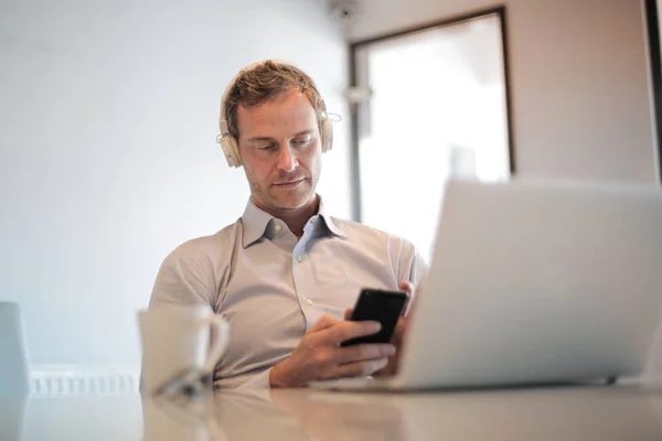 商人与耳机前的笔记本电脑阅读他的消息在手机上 — 图库照片