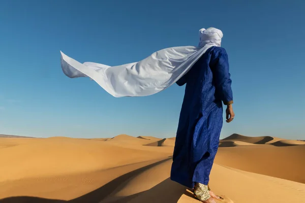 Traditional dressed Moroccan man with turban stands on a sand dune in the Sahara desert.