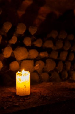 Karanlık bir mahzende yakacak odun yığınıyla yanan beyaz mum sıcak ve atmosferik bir manzara yaratıyor.