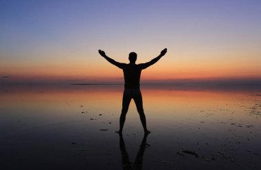 Adam kıyıda suyun içinde duruyor ve elleri günbatımının arka planında.