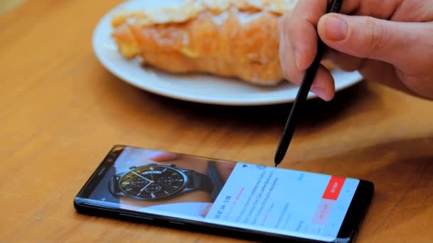 Ο άνθρωπος επιλέγει ένα ρολόι ως δώρο σε μια οθόνη smartphone σε ένα ηλεκτρονικό κατάστημα βρίσκεται σε ένα καφέ — Αρχείο Βίντεο