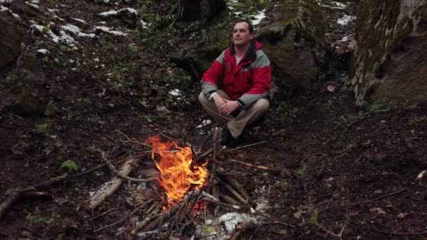 Чоловік середнього віку сидить біля багаття в лісі — стокове відео