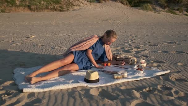 Jonge vrouw die tijdschrift leest tijdens picknick op het strand — Stockvideo