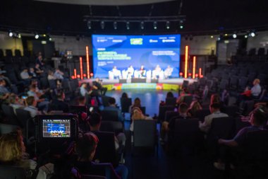 Sahnede profesyonellerin, katılımcıların oturduğu, hareketli ışıklandırmalı bir panelin olduğu bir konferans..