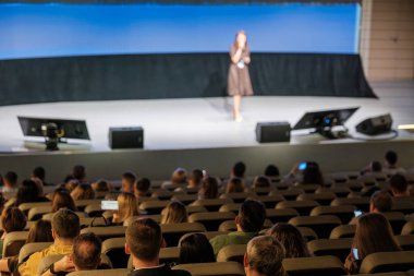 Konuşmacı, iletişim üzerine odaklanarak geniş bir konferans ortamında sahneden seyirciye sunum yapıyor..