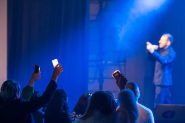 Seyirci çakmak ve telefon tutarken, adam sahnede mavi ışıkla aydınlatılmış bir şekilde şarkı söylüyor.