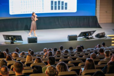 Hoparlör, modern oditoryum ortamında oturan seyircilere sahnede analitik verileri sunuyor.