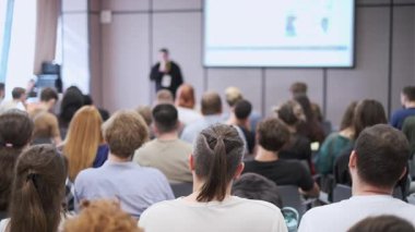 Bir grup birey, konferans salonunda sunum yapan bir konuşmacıyı dikkatle dinliyor..