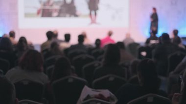 Geniş bir grup, iyi aydınlatılmış bir konferans odasında oturmuş sahnede bir konuşmacının görsel içeriği hakkında konuşmasını dinliyor..