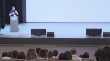 Seyirciler, bir etkinlik sırasında sahnede fikirleri sunan bir konuşmacıyı dikkatle dinlerler..