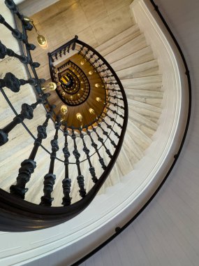 Yukarıdan görünen sarmal merdiven siyah metal balüstrad sıcak ampuller dairesel hareket derinliği ve klasik iç tasarım içeren zarif mimari detay.