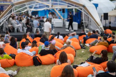 Kalabalık, sunucu barınak altında konuşurken turuncu fasülye torbalarının üzerinde rahatlıyor. Oturma ekranları canlı, çeşitli katılımcılar tartışmaya ve dinlemeye katılıyor..