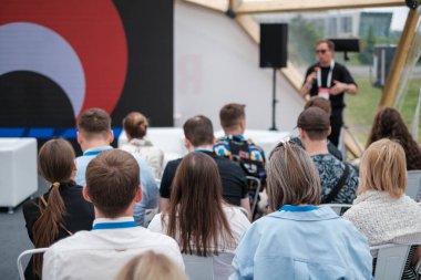 Seyirciler dışarıda oturmuş büyük kırmızı arka plan ve doğal ışıkla sahnede hoparlörleri dinliyor.