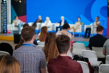 Çeşitli konuşmacılarla panel tartışma sahnesi ve renkli konferans arka planında özenli izleyiciler