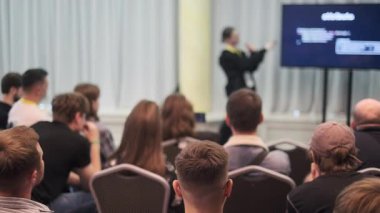 Bir konuşmacı, profesyonel bir konferans ortamında oturan seyircilere sunum yapar..