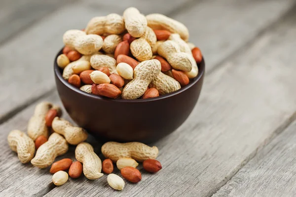 Roasted peanuts in their shells and peeled in a brown cup, against a gray wooden table. Organic vegetarian protein, macro.