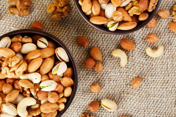 A mixture of cashew nuts, almond nuts, pistachios, hazelnuts and walnuts in a wooden cup against the background of burlap fabric. Nuts as structure and background, macro. Two cups of nuts.