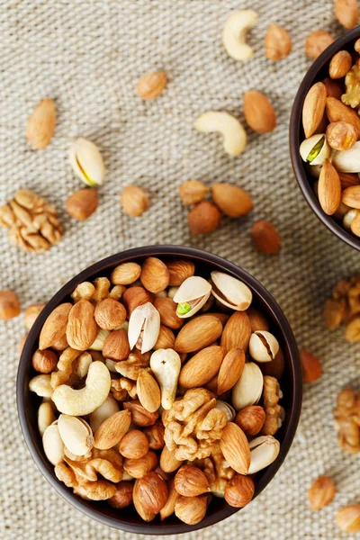 A mixture of cashew nuts, almond nuts, pistachios, hazelnuts and walnuts in a wooden cup against the background of burlap fabric. Nuts as structure and background, macro. Two cups of nuts.