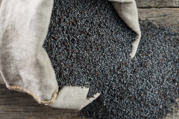 Poppy seeds in a burlap bag on a vintage wooden gray background. In country style. The tasty and useful seeds rich with protein and oils. It is poured out of a bag. Calcium content leader