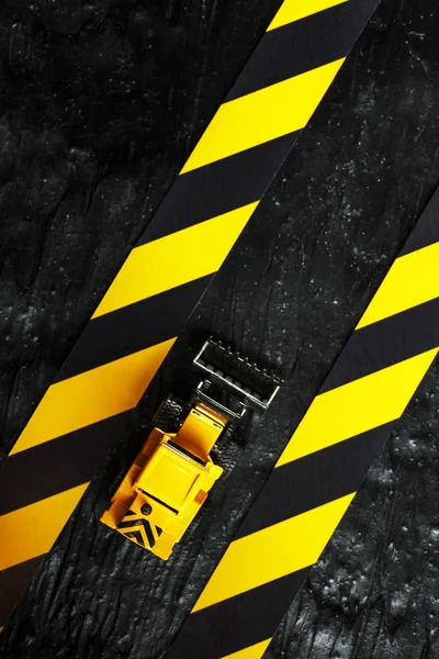 Yellow toy bulldozer on a black background texture. Black and yellow fence tape against the background of the CONCEPT