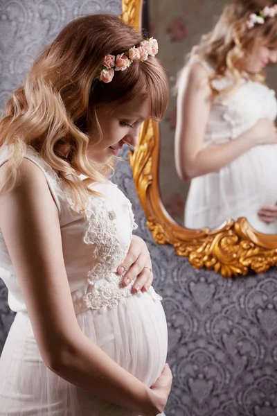 Portrait of a beautiful, young, blond long-haired pregnant mother in a white vintage peignoir, with a floral vink on her head