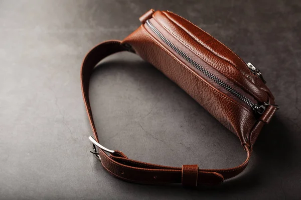 A brown belt bag made of textured brown leather on a black stone background. Elegant fanny pack brown bag with a zipper