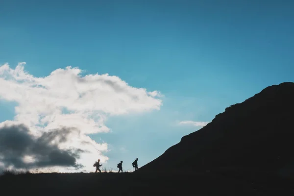 Silhouettes of travelers-tourists climbing uphill against the background of clouds and blue sky